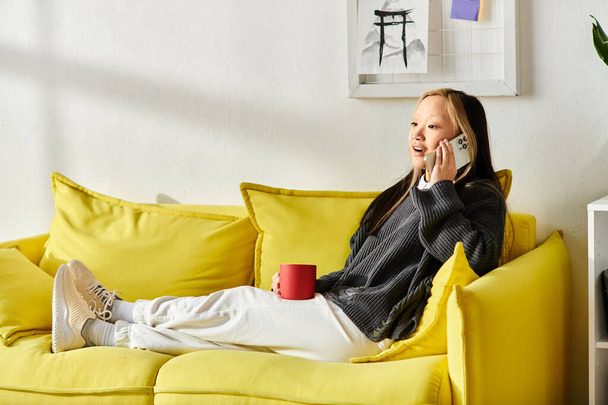 Une jeune femme asiatique est confortablement assise sur un canapé jaune, engagée dans une conversation animée sur son téléphone portable. - Photo, image