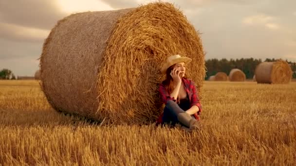Mulher agricultor na bota de borracha sentado em fardo de palha redonda e falando por telefone celular no campo de trigo colhido. Negócios na agricultura. Agricultor no campo em botas de borracha. Fardo redondo, solo fértil - Filmagem, Vídeo