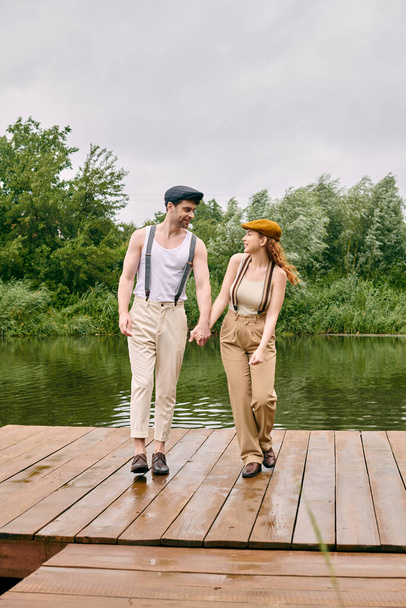 Un hombre y una mujer, vestidos casualmente, caminan de la mano a través de un puente de madera rústico en un exuberante entorno de parque verde. - Foto, Imagen