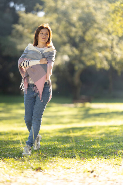 Een vrouw met een veelkleurige sjaal loopt in een park. Het park is gevuld met bomen en gras, en er is een bankje in de buurt. De vrouw geniet van haar wandeling en de vredige omgeving - Foto, afbeelding