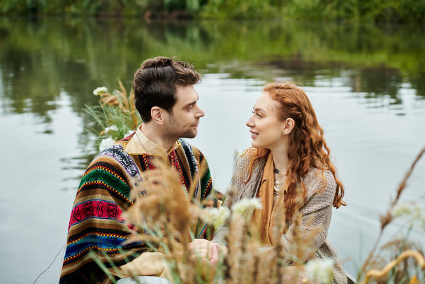 Um homem e uma mulher de traje boho relaxar por um corpo sereno de água, desfrutando de uma data romântica em um cenário exuberante parque verde. - Foto, Imagem