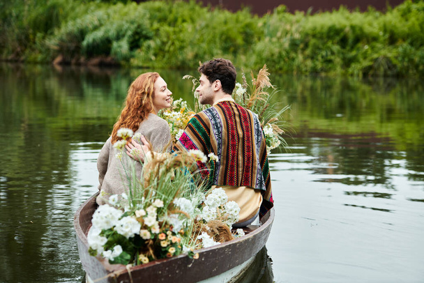 Una coppia romantica, vestita con abiti in stile boho, naviga tranquillamente in una barca ornata di fiori in un lussureggiante parco verde. - Foto, immagini