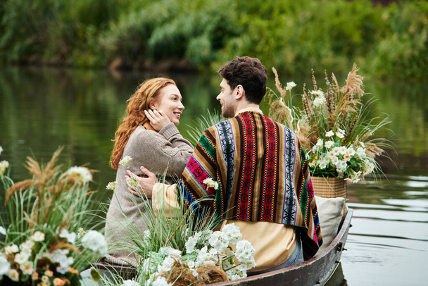 A couple in boho clothing navigate a boat filled with vibrant flowers in a serene green park setting. - Photo, Image