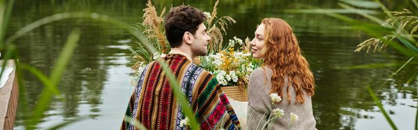 Egy elegáns pár boho öltözékben áll egy békés víztest mellett egy zöld parkban, élvezve egy romantikus pillanatot együtt.. - Fotó, kép