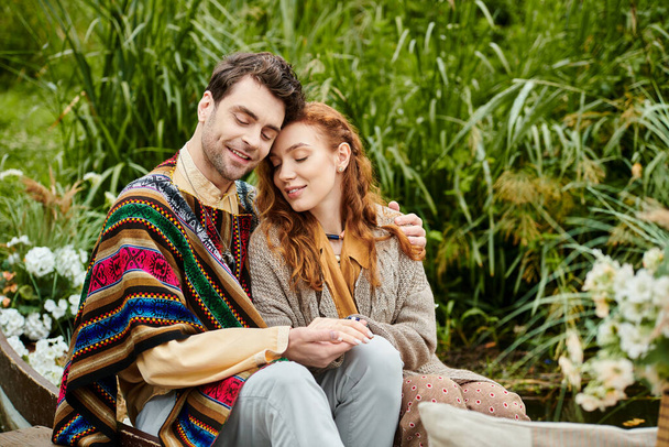 Um homem e uma mulher, ambos vestidos com roupas de estilo boho, sentam-se juntos em um parque verde, compartilhando um momento romântico.. - Foto, Imagem