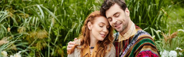Een man en vrouw, gekleed in boho stijl kleren, dicht bij elkaar in een weelderig groen park, delen een romantisch moment. - Foto, afbeelding