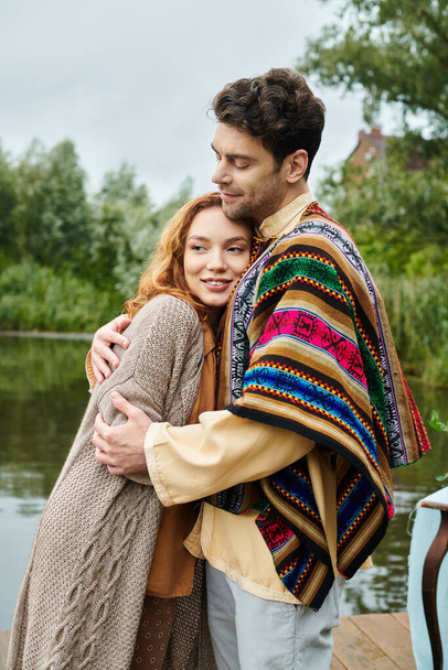 Ein Mann und eine Frau in Boho-Kleidung stehen neben einem ruhigen Gewässer in einer wunderschönen Parklandschaft. - Foto, Bild