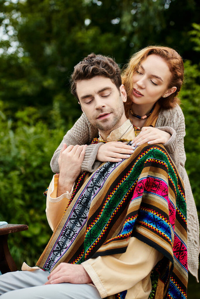 Un uomo tiene teneramente una donna avvolta in una coperta, i loro vestiti in stile boho che si fondono nel lussureggiante sfondo verde di un parco. - Foto, immagini