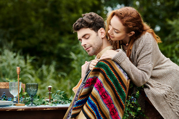 Egy boho ruhás férfi és nő ül egy asztalnál takarókkal borítva egy zöld parkban, élvezve egy hangulatos és romantikus randit.. - Fotó, kép