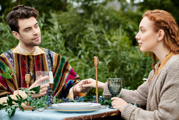 Un homme et une femme en tenue boho assis à une table dans un parc verdoyant, profitant d'un rendez-vous romantique. - Photo, image