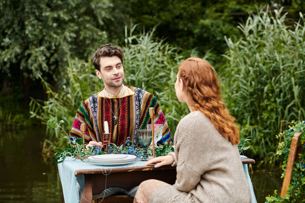 Un hombre y una mujer en ropa de estilo boho se sientan juntos en una mesa en un parque verde, disfrutando de una cita romántica junto a un tranquilo estanque. - Foto, Imagen