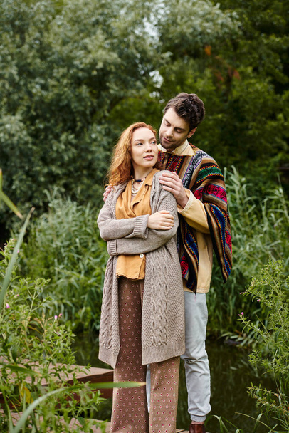Egy boho ruhás férfi és nő áll közel egymáshoz egy fahídon egy buja zöld parkban, megosztva egy gyengéd pillanatot.. - Fotó, kép