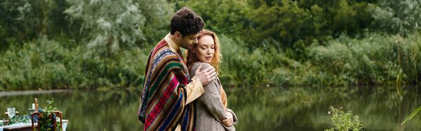 A man and woman, dressed in boho style clothes, stand next to each other near a tranquil body of water in a green park. - Photo, Image
