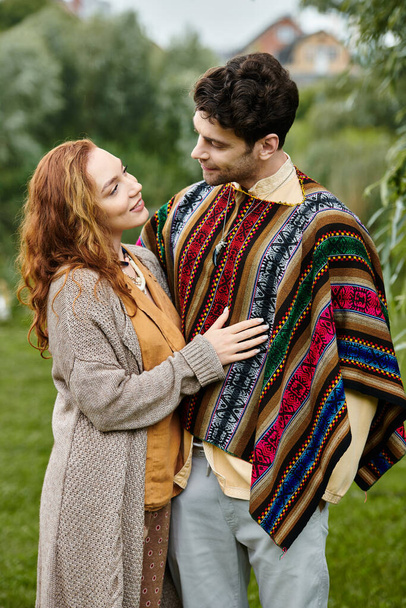 Un uomo in stile boho si trova accanto a una donna avvolta in una coperta in un sereno parco verde. - Foto, immagini