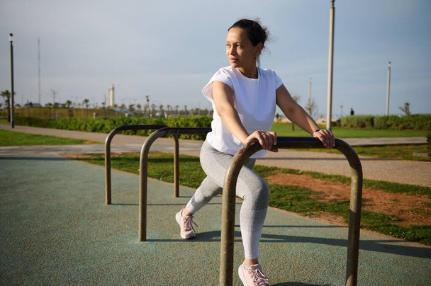 Full length female athlete exercising with crossbar on the urban sportsground. Happy determined young sportswoman enjoys her morning workout outdoors. People. Fitness. Active healthy lifestyle concept - Photo, Image