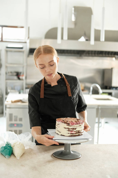 The process of preparing a cake in a professional kitchen in a pastry shop. A young female red haired pastry chef who spreads cream over a cake on a rotating stand. - Φωτογραφία, εικόνα