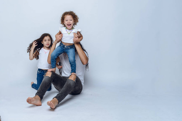 young Latino dad with long hair and glasses, sitting on floor playing with his two young daughters with curly hair, wearing white t-shirt and jean pants, studio photo with white background, copy space - Foto, afbeelding