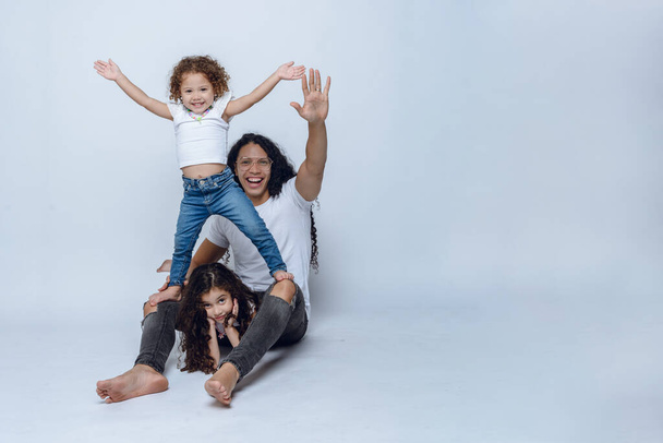 young Latino dad with long hair and glasses, sitting on floor playing with his two young daughters with curly hair, wearing white t-shirt and jean pants, studio photo with white background, copy space - Фото, изображение