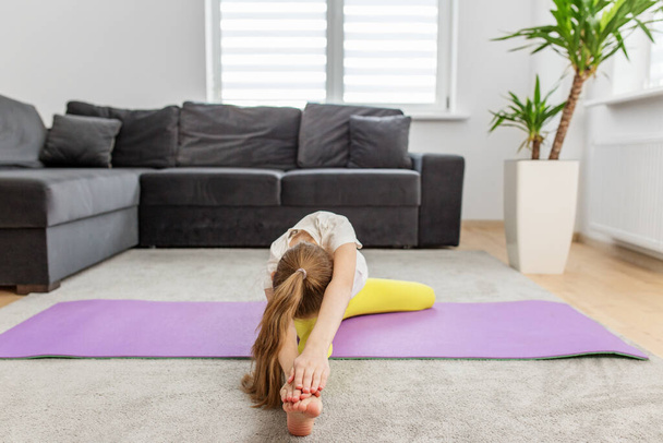 Preteen child in forward bend stretch on purple yoga mat, focusing on flexibility in home environment. - Photo, Image