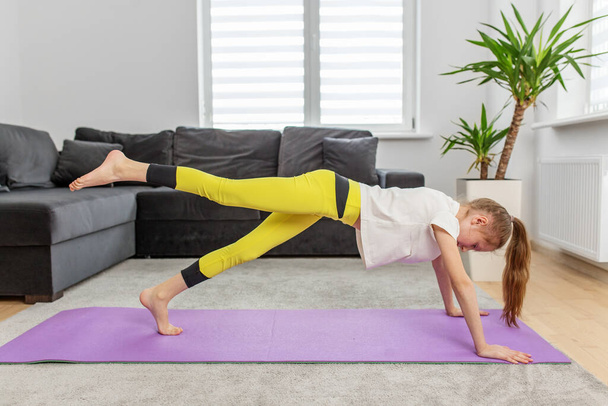 Фокусована дівчина-підліток тримає рівновагу позу під час сеансу йоги на фіолетовому килимку в сучасному домашньому обстановці. - Фото, зображення