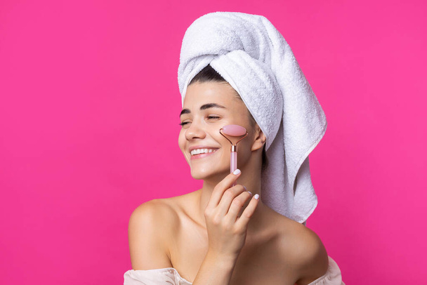 Een mooi vrolijk aantrekkelijk meisje met een handdoek op haar hoofd houdt een cosmetische roller in de buurt van haar gezicht tegen een roze achtergrond. - Foto, afbeelding