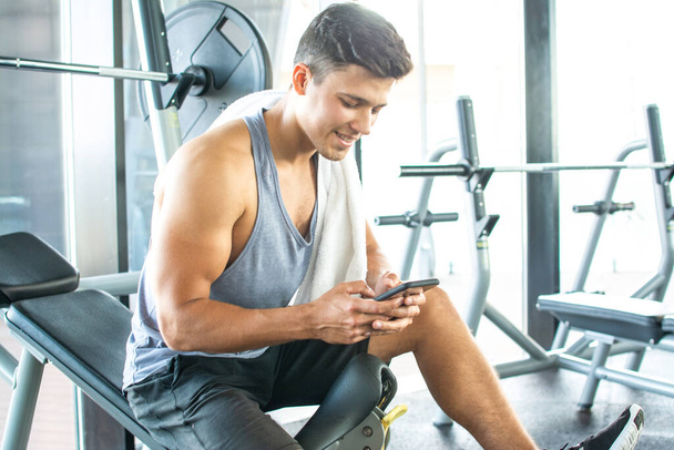 Handsome young man resting on weight bench and using phone after training at gym - Photo, Image