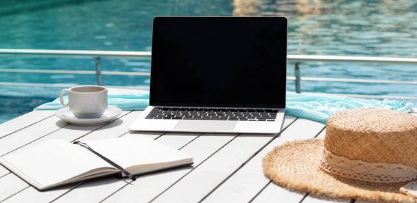 Alternative workplace at resort poolside. Laptop, notepad, cup of coffee and hat on white table isolated on blue background of pool water. Blank computer screen, copy space for text or design. Banner - Photo, Image