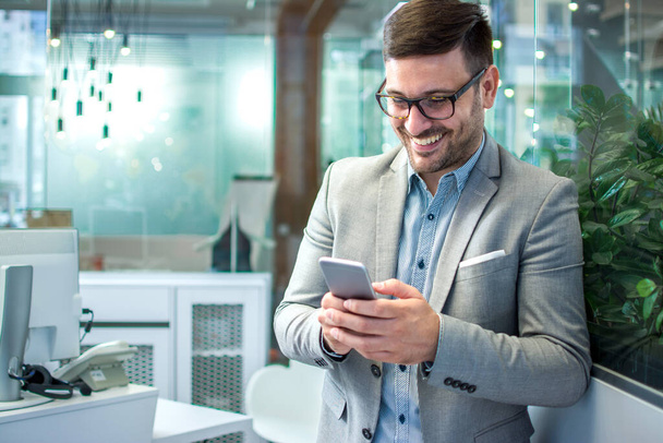 A sikeres üzletember portréja szemüvegben és formális ruházatban mosolyog, miközben üzenetet olvas és gépel a vállalati irodában. - Fotó, kép