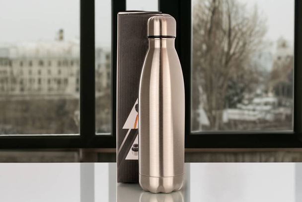 One metal thermos bottle with a paper box is on the table, close-up, against the background of the window. - Photo, Image