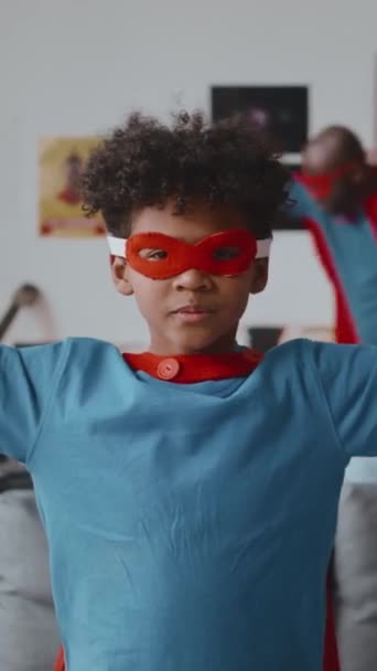 Vertical portrait shot of little curly haired African American boy in superhero cape and mask showing biceps and posing on camera while father running behind in living room - Footage, Video