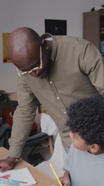 Imagen vertical del niño negro dibujando en el escritorio en la habitación de los niños mientras el padre camina hacia él y alaba - Metraje, vídeo