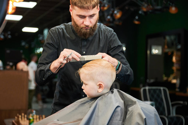 Side view of cute boy getting stylish haircut. Hairdresser in process. Little client first time in beauty salon. - Photo, Image