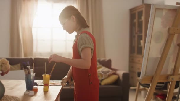 Średni materiał filmowy młodej utalentowanej dziewczyny myjącej pędzel w wodzie i kontynuującej malowanie na płótnie sztucznym ramieniem będąc w domu samotnie w słoneczny dzień - Materiał filmowy, wideo
