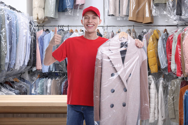 Happy courier holding coat in plastic bag and showing thumbs up in dry-cleaning - Photo, Image