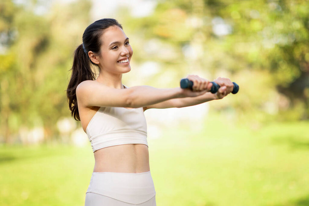Stralende glimlachende blanke jonge vrouw atleet met een stralende glimlach oefeningen met de hand gewichten in een weelderig groen park, waaruit blijkt een toewijding aan haar gezondheid en fitness, buiten - Foto, afbeelding
