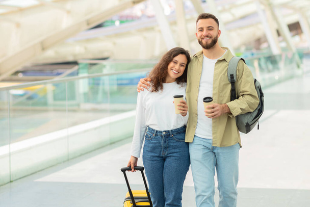 Un hombre y una mujer sonrientes sosteniendo tazas de café y equipaje en un aeropuerto, lo que sugiere que son viajeros o de vacaciones - Foto, imagen
