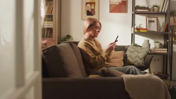 Side through-doorway footage of Caucasian girl coming to mom who sitting on sofa with phone and hugging her from behind - Footage, Video