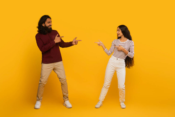 Hombre y mujer con atuendo casual se apuntan juguetonamente el uno al otro sobre un fondo amarillo vivo - Foto, imagen