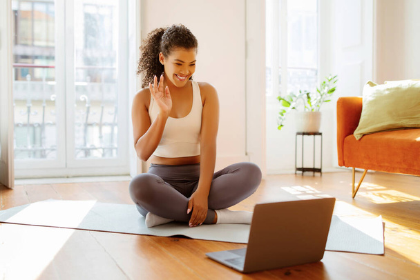 Sporty Brazilian woman in sportswear waves hello to laptop while practicing online yoga session at home, blending modern technology with active wellness and workout routine - Foto, Bild