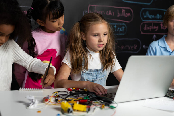 Inteligentna dziewczyna w białym śliniaczku uczy się kodowania technologii robotyki przy użyciu laptopa w klasie STEM. Uczeń w niebieskiej koszuli stara się edukować płytę główną, podczas gdy inteligentne uczennice czytają kod. Erudycja. - Zdjęcie, obraz