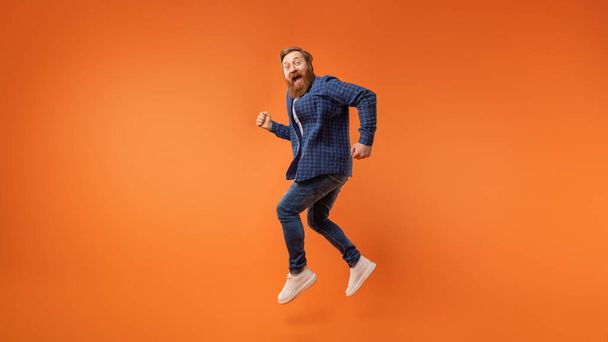 Excited redhaired bearded guy wearing casual outfit jumping and shouting joyfully over orange studio backdrop, expressing positive energetic vibe, having fun. Full length, panorama with copy space - Photo, Image