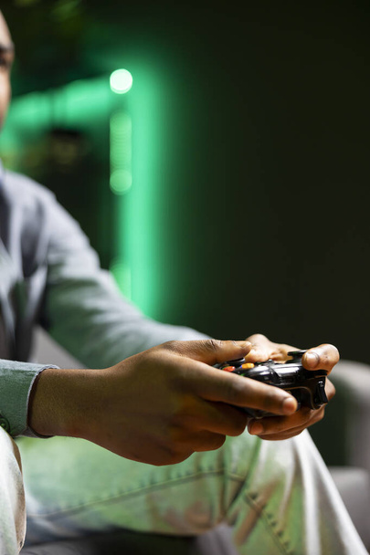 Hráč na gauči pomocí ovladače a sluchátek k přehrávání videoher v neonově osvětleném bytě, zblízka. Hráč v RGB osvětlené obývací pokoj pomocí high-tech herní konzole joystick - Fotografie, Obrázek