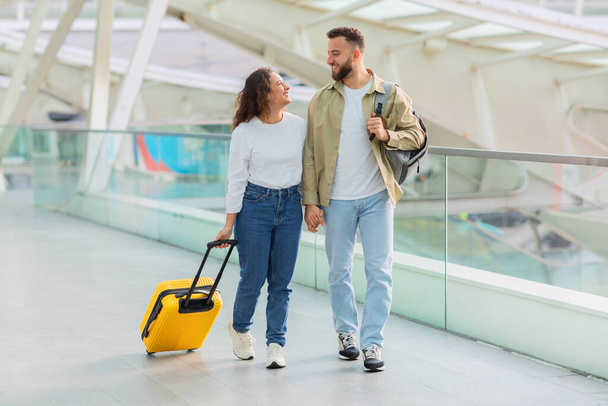Genç bir çift, günümüzün cam duvarlı havaalanı terminalinde, seyahat ve birlikteliği simgeleyen sarı bir bavulla gelişigüzel yürüyor. - Fotoğraf, Görsel
