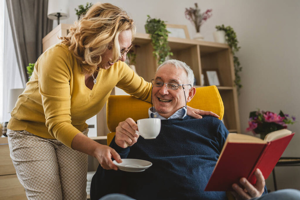 Hombre mayor leyó el libro mientras su esposa le traía una taza de café en casa. - Foto, imagen