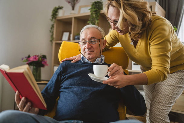 Homme âgé lire livre tandis que sa femme lui apporter une tasse de café à la maison - Photo, image