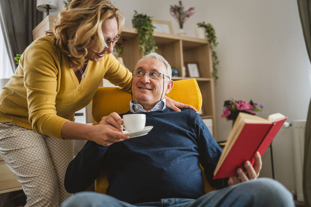 Yaşlı adam kitap okurken karısı ona eve bir fincan kahve getiriyor. - Fotoğraf, Görsel