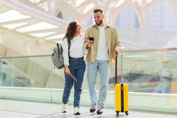Ευτυχισμένο ζευγάρι βόλτα με μια κίτρινη βαλίτσα, ενώ ασχολούνται με τη συζήτηση σε ένα smartphone - Φωτογραφία, εικόνα