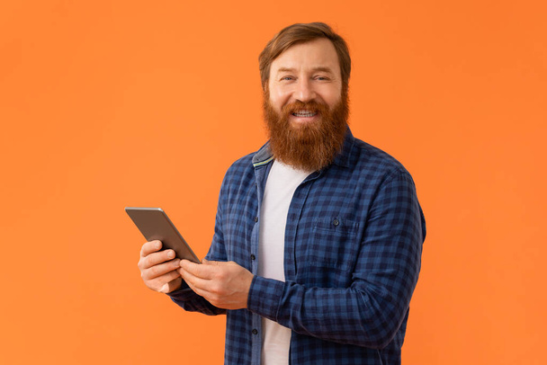 Vidám vörös hajú szakállas férfi alkalmi gazdaság digitális tabletta számítógép, pózol eszköz és webszörfözés narancssárga stúdió háttér. Az internetes szabadidő és szórakozás fogalma - Fotó, kép