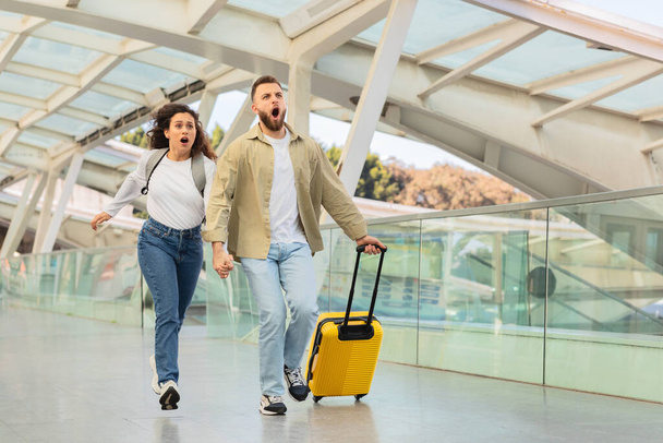 Dos personas en atuendo casual están corriendo con expresiones de pánico, posiblemente tarde para un vuelo, en un aeropuerto arquitectónicamente moderno con una maleta amarilla - Foto, imagen