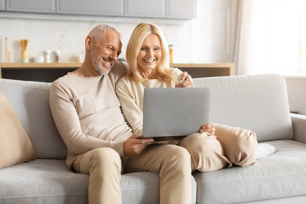 Alegre pareja madura divirtiéndose mientras se sienta e interactúa con una computadora portátil en el sofá - Foto, imagen